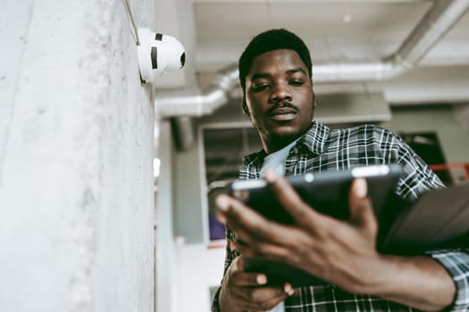A young man stands in an office, focused on a tablet while checking surveillance cameras mounted on the wall. The industrial-style space is well-lit and contemporary.
