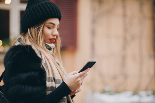 Woman using cell phone in cold weather outside in city urban setting. Technology, winter and holiday concept. Girl Enjoying Winter Holiday Season, social networking, using app.