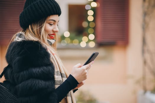 Pretty girl walking against a wall with Blurred Christmas Lights on background, she has bright makeup and red lipstick. Woman using cell phone in cold weather outside in city urban setting.