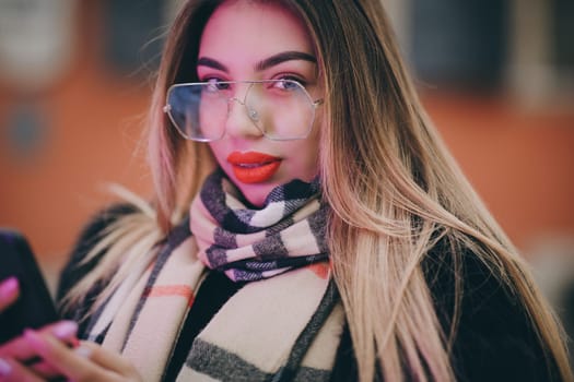 Portrait woman with glasses look at camera poses in city center Beautiful hair attractive smile girl. Close shot on hands using phone during winter time. Technology, winter and holiday concept.