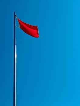 A red flag flutters in the wind in the blue sky by the sea during the day. Danger sign in the form of a red flag.