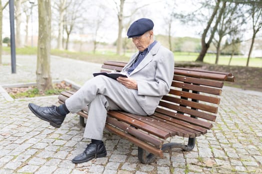 Senior man wearing earphones is taking notes in his notebook while sitting on a bench in a park