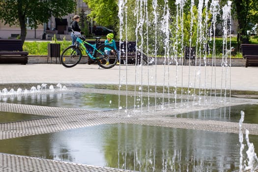 A person skillfully riding a bicycle in front of a beautifully designed fountain, enjoying the refreshing sight and sound of water splashing all around