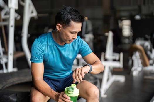 A focused man in fitness attire sits on gym equipment, checking his smartwatch while holding a protein shake, emphasizing fitness and health.