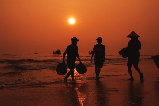 Silhouettes of Vietnamese people on the beach by sea with ships in fish market in fishing village in morning at sunrise in Vietnam. Tam Tien, Quang Nam province, Vietnam - April 10, 2025