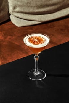 Cocktail glass containing a refreshing beverage, adorned with a dried orange slice, sits on a sleek black table in a dimly lit bar with a soft, inviting ambiance