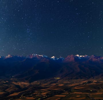 A breathtaking starry night sky illuminates the rugged, snow-capped mountains of Kyrgyzstan in summer, with a serene valley and scattered village lights below.