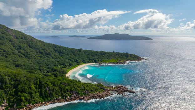 Waves gently roll onto a white sandy beach, bordered by lush green vegetation. La Digue, Seychelles.