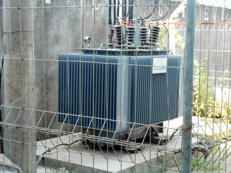 High Voltage Transformer Enclosed Behind Metal Fencing in an Industrial Setting, An image depicting a high-voltage electrical transformer protected by a secure fence in an industrial environment, emphasizing industrial infrastructure and safety measures for operational energy equipment.