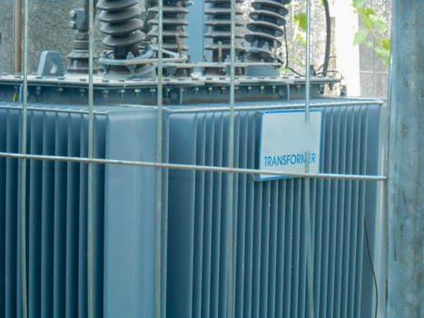 High Voltage Transformer Enclosed Behind Metal Fencing in an Industrial Setting, An image depicting a high-voltage electrical transformer protected by a secure fence in an industrial environment, emphasizing industrial infrastructure and safety measures for operational energy equipment.