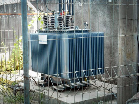 High Voltage Transformer Enclosed Behind Metal Fencing in an Industrial Setting, An image depicting a high-voltage electrical transformer protected by a secure fence in an industrial environment, emphasizing industrial infrastructure and safety measures for operational energy equipment.