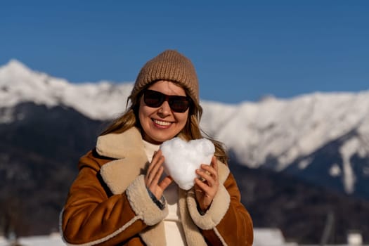 Smiling woman in sunglasses and brown coat holds a snow heart, mountains in the background. Sunny winter day.