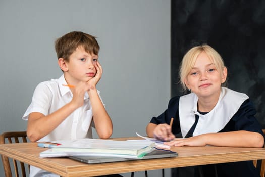 A boy and girl sit at a desk. The boy rests his head on his hand, seemingly bored, while the girl writes in a notebook.