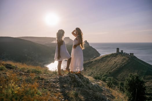 Two women are standing on a hill overlooking the ocean. They are holding hands and looking out at the water. The scene is peaceful and serene, with the sun shining brightly in the background