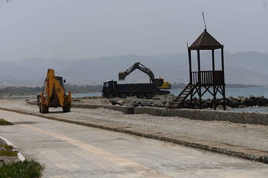 Heavy-duty yellow excavator loads large sea rocks into a dump truck on the Mediterranean coast of Cyprus during shoreline construction.