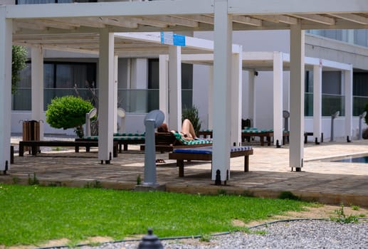 Relaxed woman sunbathes on a lounge chair near a turquoise pool in a residential complex in Cyprus on a bright sunny day.