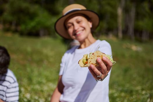 A woman, happily enjoying time outdoors, holds out a taco during a picnic. The lush green setting and sunny atmosphere emphasize leisure, togetherness, and joyous outdoor dining.