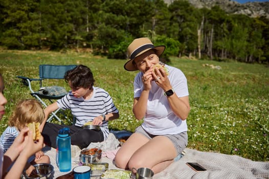 A joyful mother shares a picnic outdoors with her children, enjoying tacos in a serene mountain meadow. Togetherness, fun, and connection characterize this healthy, active moment in nature.