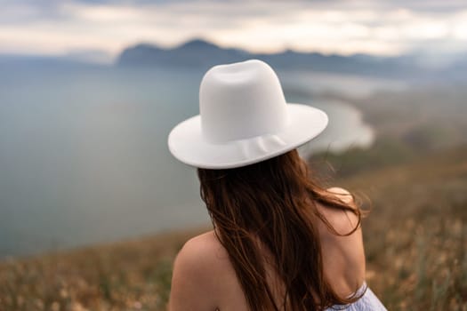 Woman in a white hat stares at the sea meeting distant hills. Back view, dry grass in foreground, soft light.