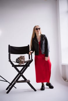 A woman, freelancer, blogger in a silk red dress on a white floor, black background. blonde owner cat of the Scottish Straight-eared breed at production studio