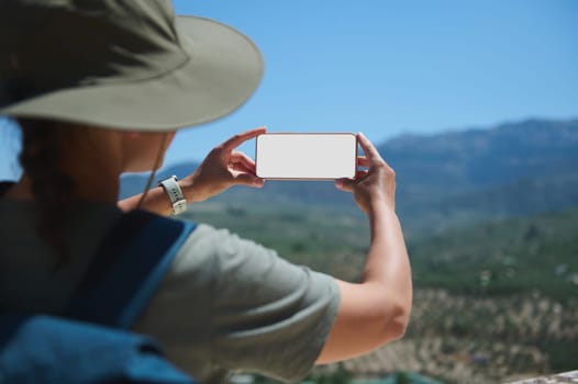 A tourist wearing a hat using a smartphone to capture stunning mountain vistas on a sunny day, showcasing nature and travel exploration.