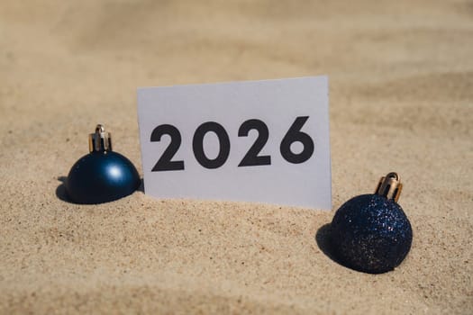 2026 text on paper greeting card on background sandy beach sun coast. Christmas balls New Year New Me decoration. Summer vacation decor. Holiday concept calendar date postcard. Getting away Travel Business concept