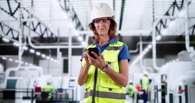 Female Factory Worker Inspecting Automated Production Line While Using Smartphone.