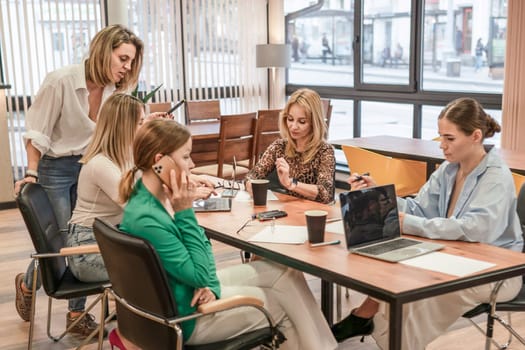 Five women meet at a table indoors, using laptops, taking notes, and talking on a phone.