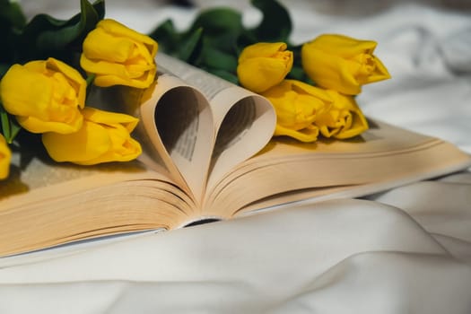 Book pages in shape of heart symbol love with bouquet of pink tulips on messy bed. Romantic morning scene. Greeting card Holiday flowers springtime atmosphere