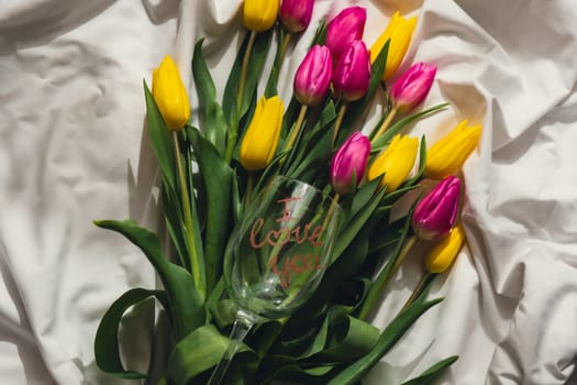 Wine glass with words I LOVE YOU Fresh bouquet of colorful tulip flowers on bed. Gift present idea for holiday birthday 8 March Mother's Day Valentine's day Women's Day. Copy space