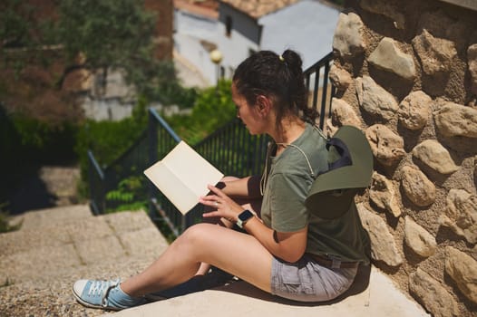 A woman in casual attire, enjoys reading a book while seated on steps near a rustic stone wall surrounded by greenery in a tranquil outdoor environment.