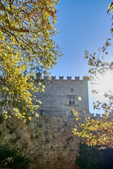 Glimpse the historical grandeur of Grossmeisterpalast in Rhodes-Stadt, with its aged stone facade appearing behind a curtain of vibrant green and golden leaves against a bright sky.