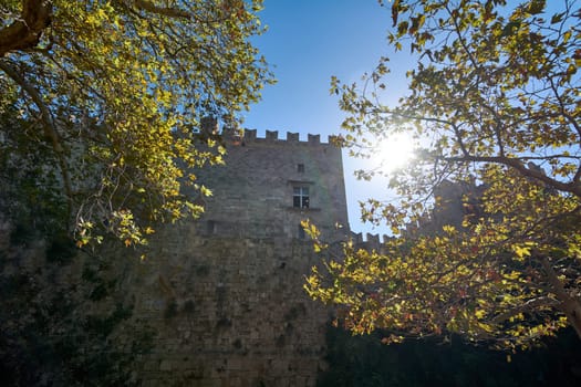 The medieval architecture of the Grossmeisterpalast stands majestically, its imposing stone walls and classic windows partially veiled by the rich, autumnal canopy of nearby trees.