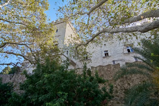 A unique perspective of the ancient Grossmeisterpalast, showcasing its sturdy medieval construction framed by the natural beauty of changing tree foliage, bathed in sunlight.