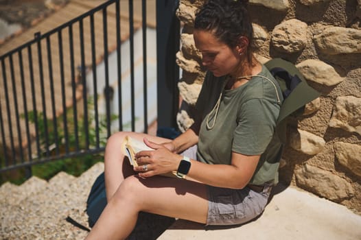 A woman enjoys a peaceful moment reading a book against a scenic medieval stone backdrop, representing active tourism and nature appreciation in a historical mountain setting.