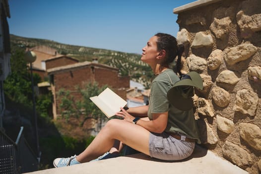 A traveler enjoying the outdoors while resting against a stone wall with an open notebook. The setting features a rustic village and picturesque greenery, embodying a peaceful and contemplative mood.