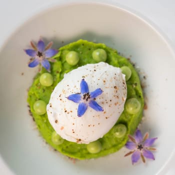 A fresh pea puree is beautifully plated, featuring a poached egg at the center and adorned with borage flowers. The dish showcases vibrant colors and textures, perfect for any spring menu.