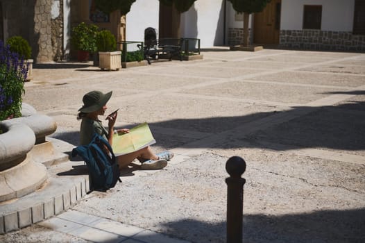 A lone traveler sitting in a historic plaza, examining a map under the bright sunlight, exudes an essence of adventure and discovery in a scenic and tranquil town.