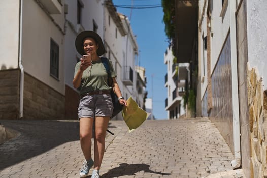 Adventurer walks in a charming Spanish cobblestone street, holding a map and phone, enjoying traditional architecture with sunny weather.