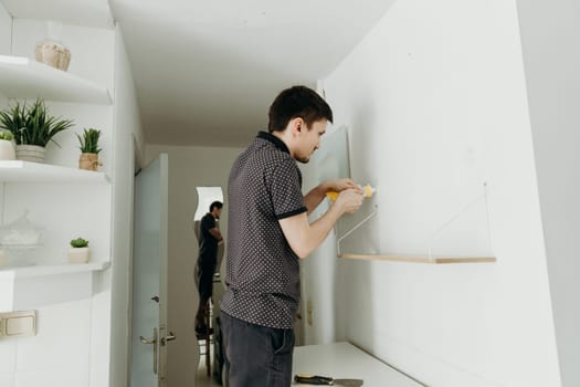 One young handsome Caucasian brunette guy stands half sideways and squeezes a special putty from a tube into a screw hole on a white wall, standing in a room with a narrow passage on a spring day, side view close-up with depth of field.