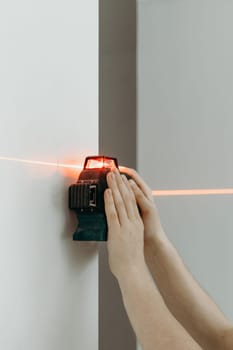 One young unrecognizable Caucasian guy holds with both hands, using a modern laser level to measure a straight line on the wall, before drilling with a drill, side view close-up.