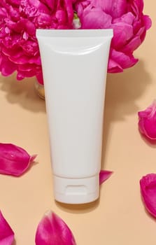 Close-up of white cosmetic tube without label surrounded by bright pink peonies and their petals on beige background.