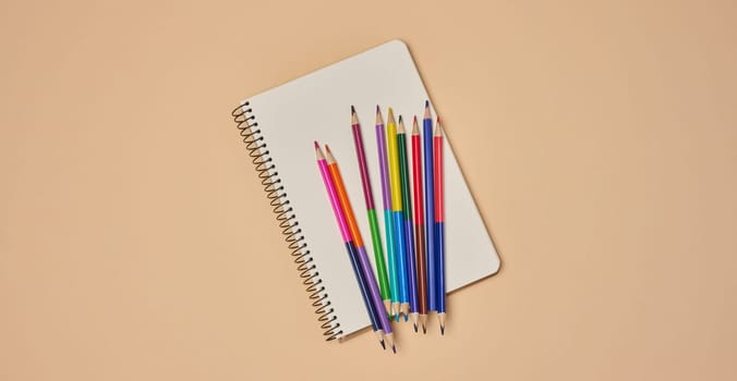 Top view of an open notebook with pastel-colored pencils on a beige background. Flat lay composition of school or office stationery supplies.