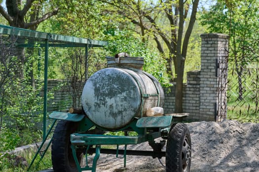 An old metal tank on a trailer, covered with traces of rust and time, stands against the background of green trees and a chain-link fence. The trailer is painted a worn green color