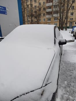Car Covered in Snow after Snowfall. High quality photo A grey car is completely covered in fresh white snow, parked outside a residential building during a winter day. Trees without leaves and more snow visible in the background.