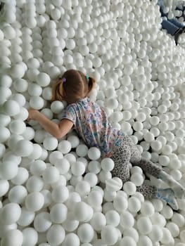 A young girl with pigtails and patterned clothing lies among many white balls in a ball pit. She is partially submerged, enjoying her time in the colorful play area.