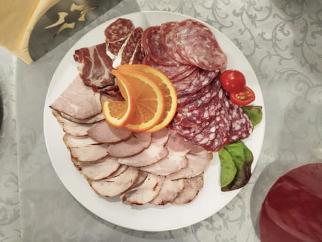 An assortment of sliced cold cuts, salami, ham, an orange slice, and tomatoes are arranged on a white plate. There is a napkin and a red lid on the patterned tablecloth.