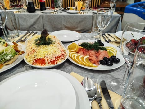 Appetizing food preparations are served on plates, including salads, fish, lemon slices, olives, and greens. Glassware and cutlery are placed on the table for dining.