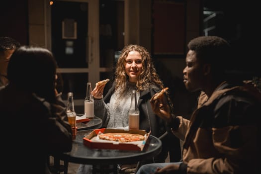 Friends gather around a table under dim lighting, sharing laughter and a pizza. Bottled drinks sit beside them as they savor their meal and enjoy each other's company.