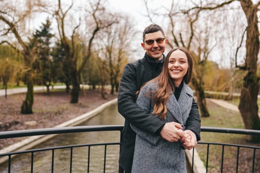 A couple shares a warm embrace on a bridge surrounded by bare trees and a calm waterway, enjoying each other's company on a chilly afternoon in early spring.
